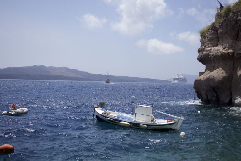 Small white and blue boat moored beside a rocky cliff, with a sailing ship and cruise ship on open water